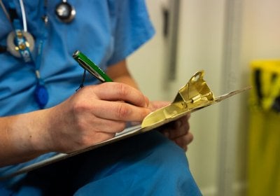Healthcare worker in blue scrubs writing notes on a clipboard in a clinical setting.