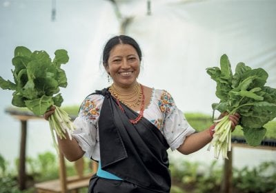 A south American women stands on a crop patch and holds up two bunches of leaves, smiling
