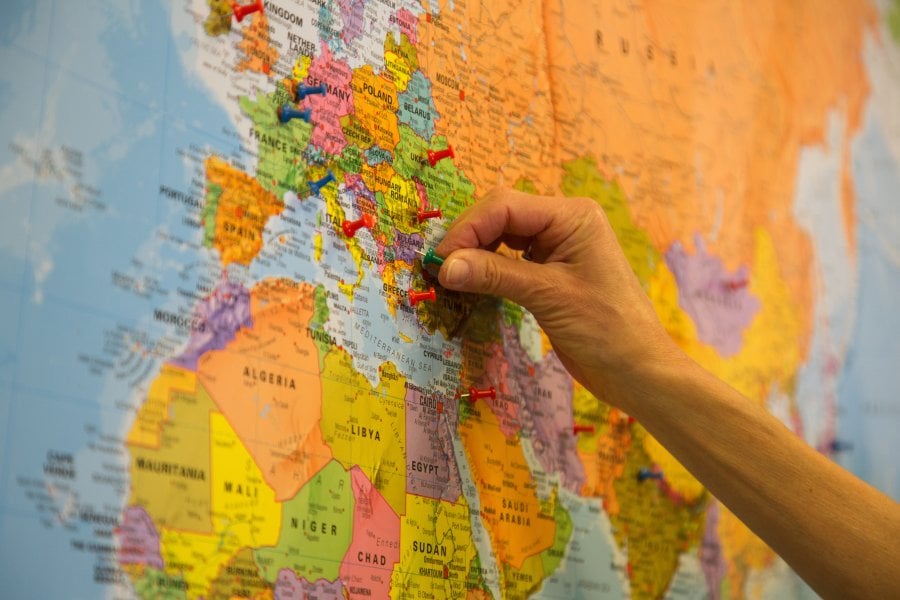 Close-up of a colorful world map focused on Europe and parts of North Africa and the Middle East, with multiple push pins marking locations. A hand is placing a green pin near southeastern Europe.