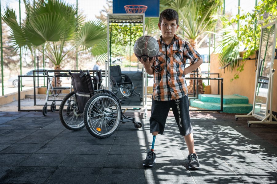 A young child with prosthetic leg standing up, holding a ball in one hand. Pictured behind is a wheelchair in the background. Photo credit: Rachel Elkind, Relief International. 
