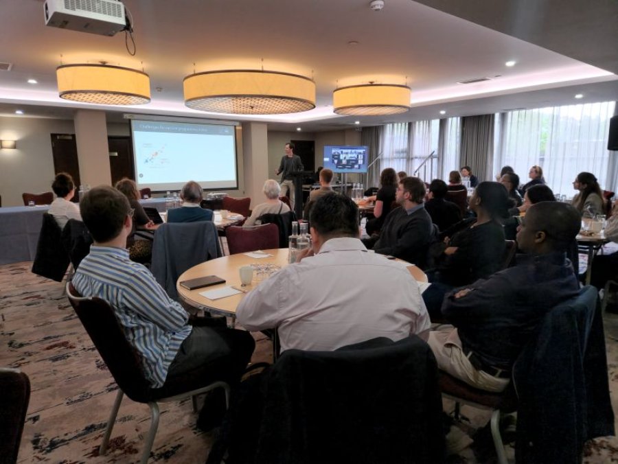 People sitting around round tables, listening to someone standing at lecturn giving presentation in hotel conference room.