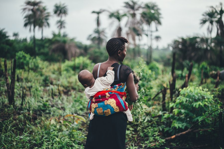 Mother carrying a young child on her back, shown from behind, in green fields in Sierra Leone