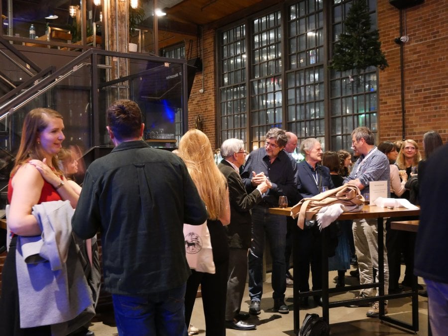 A group of people stand and talk at a networking reception inside a modern venue with exposed brick walls, high ceilings, and large industrial-style windows.