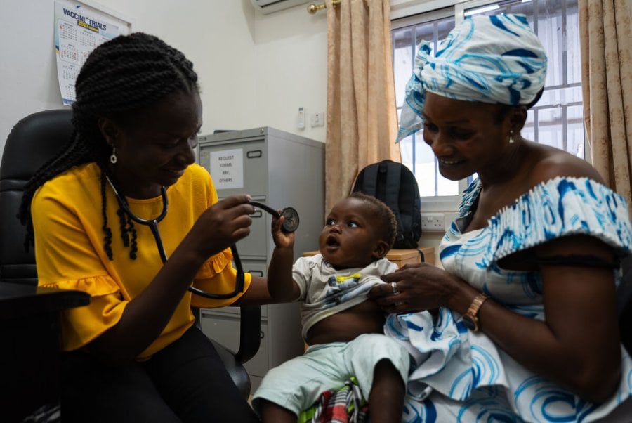Healthcare worker using a stethoscope to examine a young child seated on a caregiver’s lap in a clinic setting,