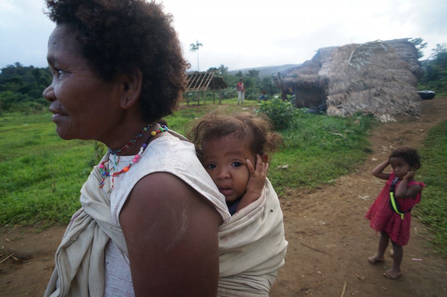 A woman carrying a baby on her back, being watched by a toddler standing in the background. 
