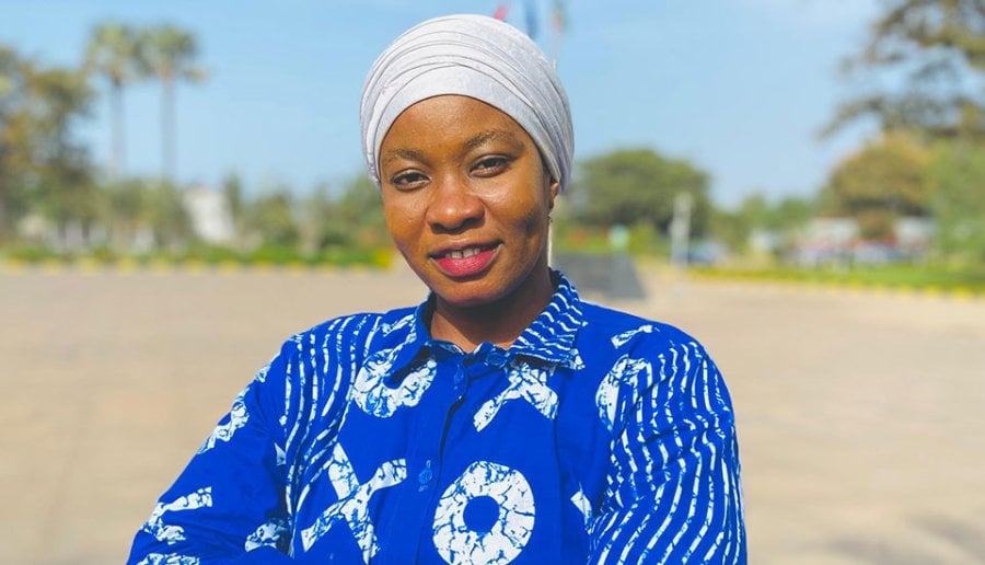 Woman with blue shirt with a white print stands arms folded with an rural landscape in the background in The Gambia.