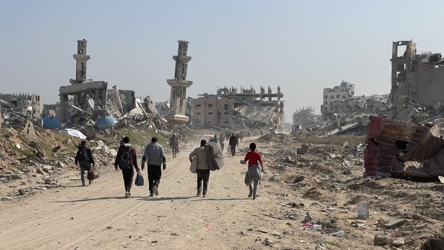 people walking along a dusty road in Gaza, carrying household items, with ruined and collapsed buildings in the background against a blue sky