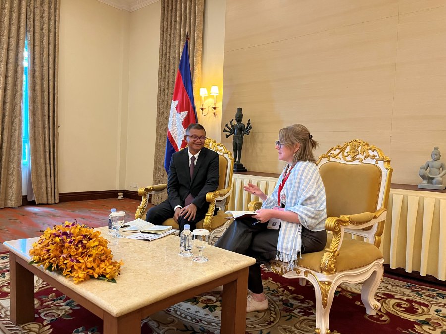 A white woman wearing a shawl and an Asian man wearing a suit sit in a formal room with a Filipino flag behind them