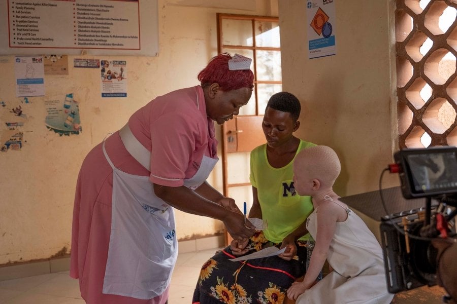 A nurse at a health facility in Luuka district, Uganda is assisting a mother of a child with albinism. She is handing over a prescription to the mother and her daughter, who is sitting next to her looks at the piece of paper. In the corner, a film camera is visible.