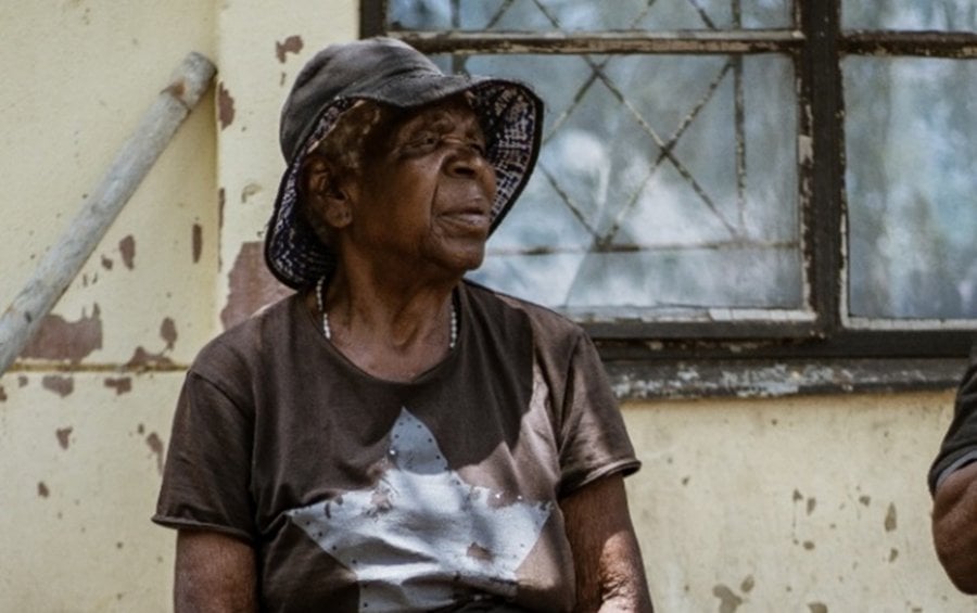 An older black woman looks into the distance. She is wearing a black floppy hat and a black t-shirt.