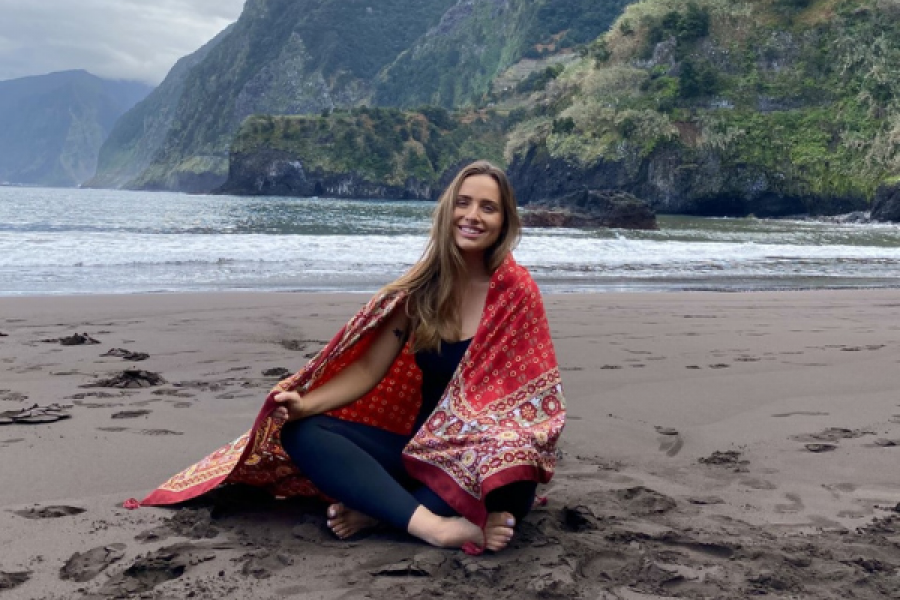 Sabrina is sat cross legged on sand with a blanket wrapped around her. In the background is a lake and mossy cliffs. 