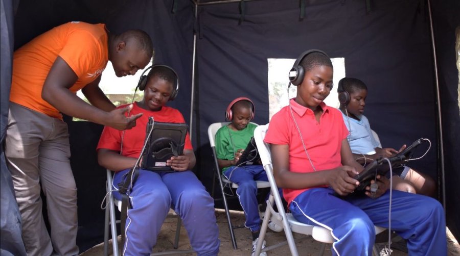 Four adolescents sit with electronic tablets and headphones completing questionnaires. A man stands beside one of the boys, supporting them on the tablet.