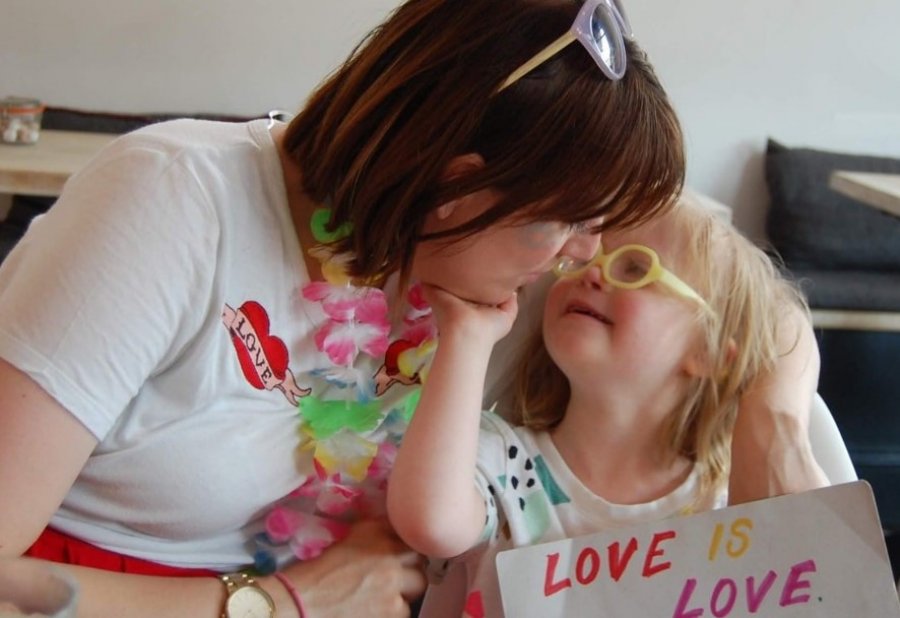 Vicky and Audrey cuddling. Child is sitting on a high chair holding a placard that says "love is love". Photo credit: Disabled Children’s Partnership 