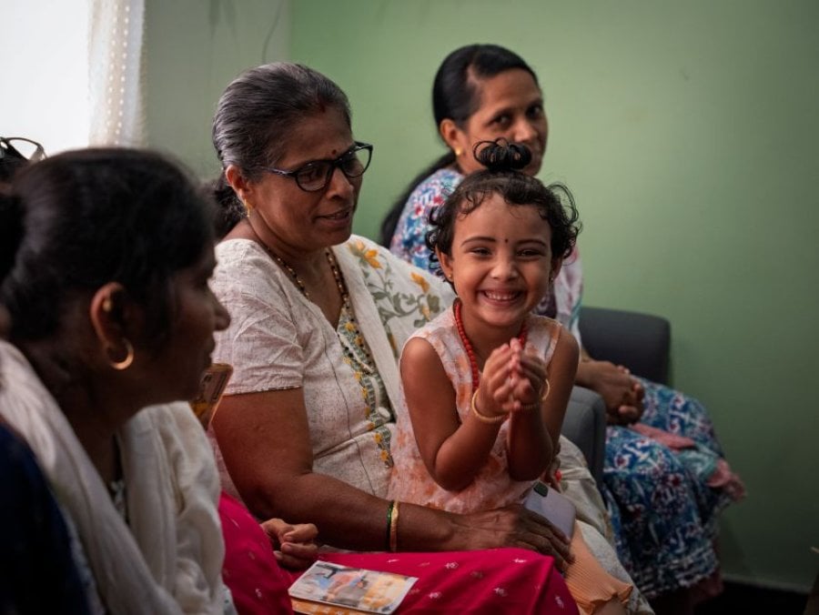A child smiles towards the camera while sitting on a woman's knee. Another woman sits behind her while a different woman sits in front of her