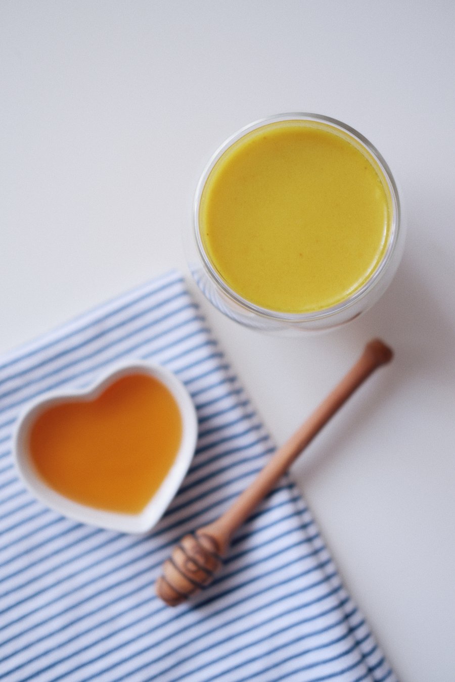 A glass containing turmeric golden milk, nearby a heart shaped dish containing honey