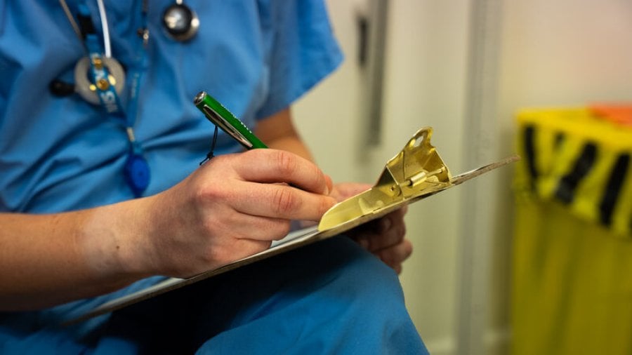 Healthcare worker in blue scrubs writing notes on a clipboard in a clinical setting.
