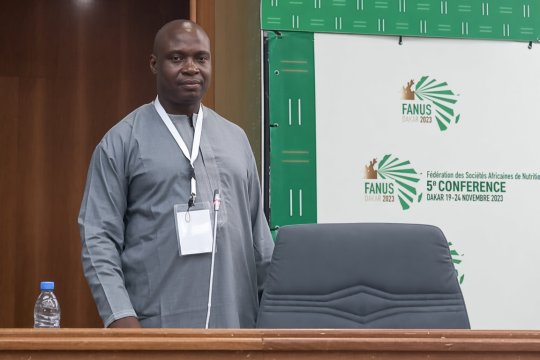 Robert Fungo dressed in a grey African robe standing in front of a panel for a conference