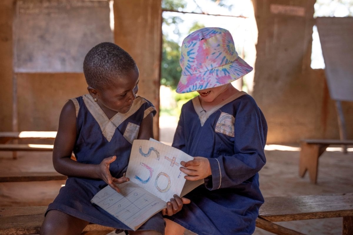 Two children sit in a classroom in Luuka, Uganda. They are holding a book and smiling. One of the children is wearing a bucket hat.