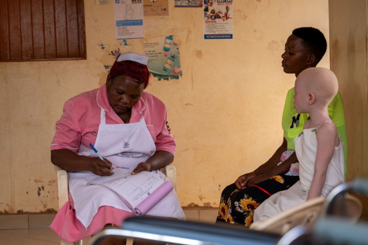 A nurse at a health facility in Luuka district, Uganda is assisting a mother of a child with albinism. She is handing over a prescription to the mother and her daughter, who is sitting next to her, looks at the nurse.