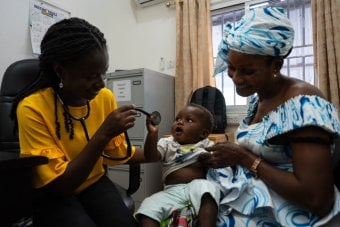 Healthcare worker using a stethoscope to examine a young child seated on a caregiver’s lap in a clinic setting,