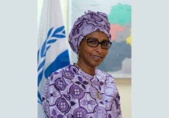 Fathiaa Abdalla wears a purple patterned headdress and robe and stands in front of a UN flag