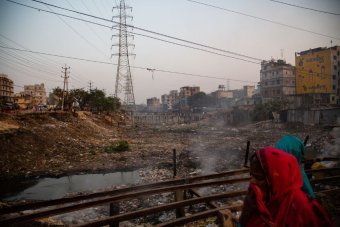 	A woman crosses a bridge spanning a dry riverbed and covers her face from the fumes of burning rubbish, Dhaka