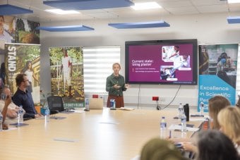 A presenter speaks during the STAND project inception meeting at MRC Gambia, showing a slide on plaster use while attendees listen around a conference table. The STAND initiative supports amputees in The Gambia.