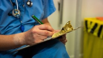 Healthcare worker in blue scrubs writing notes on a clipboard in a clinical setting.