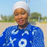 Woman with blue shirt with a white print stands arms folded with an rural landscape in the background in The Gambia.