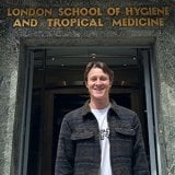 Student, Thomas, standing outside the LSHTM building entrance wearing a white t-shirt and patterned jacket.